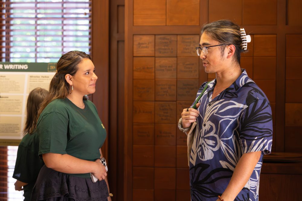 Two individuals engage in conversation indoors, one wearing a green shirt and the other in a patterned blue shirt with glasses and a hair clip.