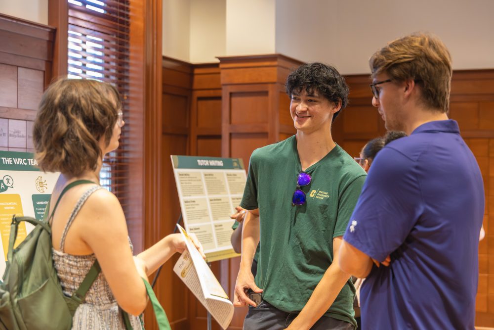 Three individuals engage in conversation near informational displays in a warmly lit room with wooden paneling.