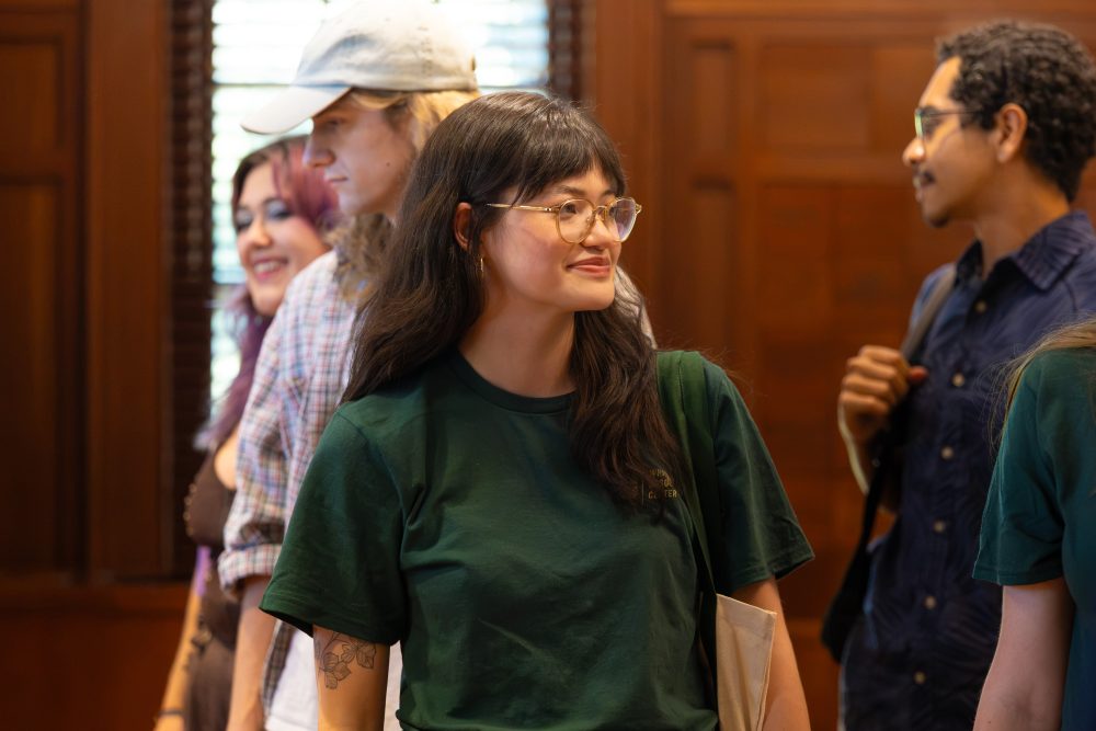 A group of people stand indoors, with one person in a green shirt and glasses smiling while looking toward another individual.