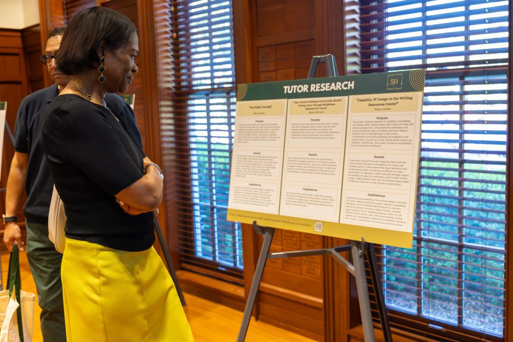 A person in a black top and yellow skirt stands before a research display board titled "Tutor Research," while another individual observes nearby.