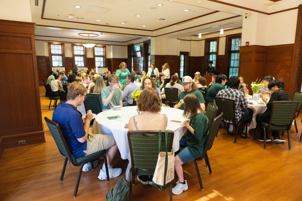 A group of people are seated at round tables in a large room with wooden floors and paneling, enjoying a meal together.