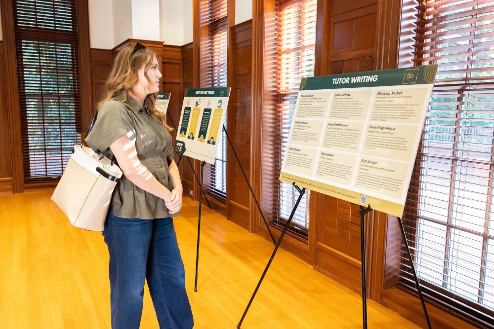 A person stands in a warmly lit room with wooden floors and windows, examining informational displays on easels.