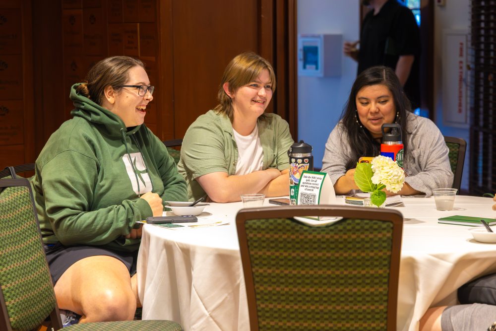 Three individuals are seated around a table, laughing and engaging in conversation during what appears to be a casual gathering.