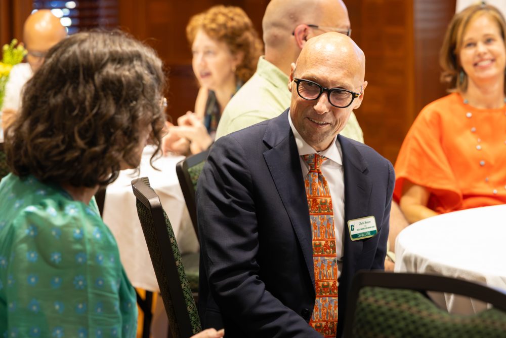 A man in a suit and patterned tie smiles while conversing with another person at a formal gathering.
