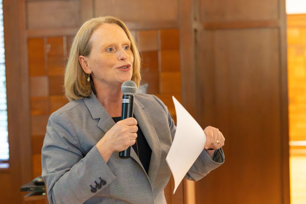 A person with blonde hair speaks into a microphone while holding a piece of paper, standing in front of a wooden-paneled background.