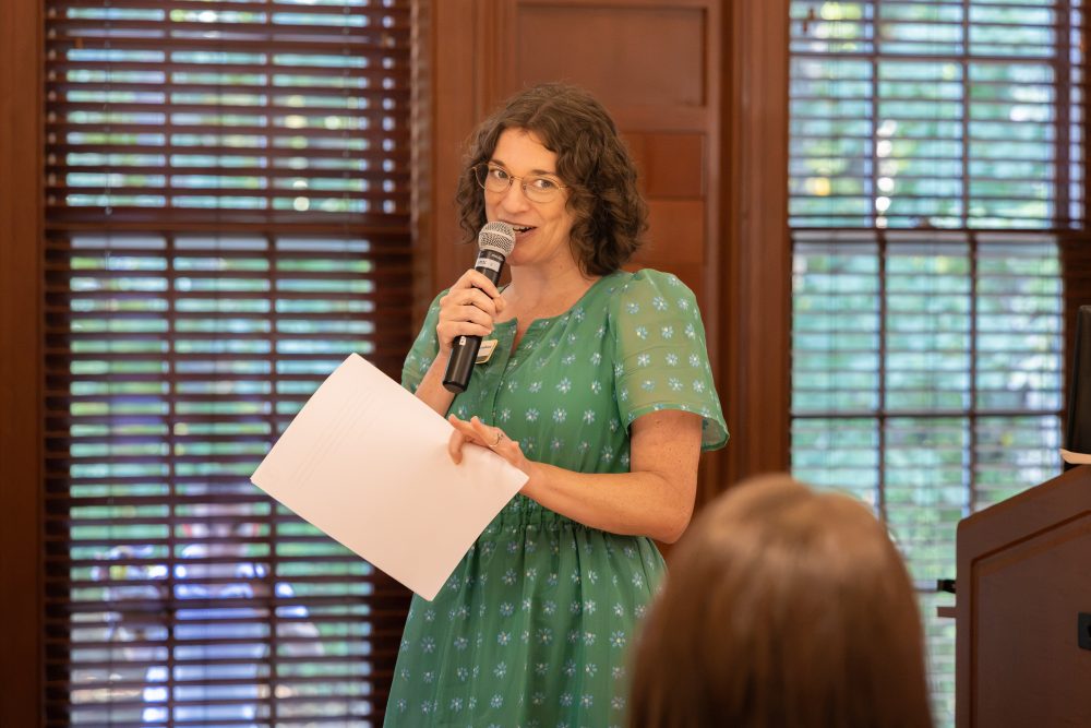 A person in a green patterned dress stands holding a microphone and papers, speaking to an audience in a room with wooden blinds.