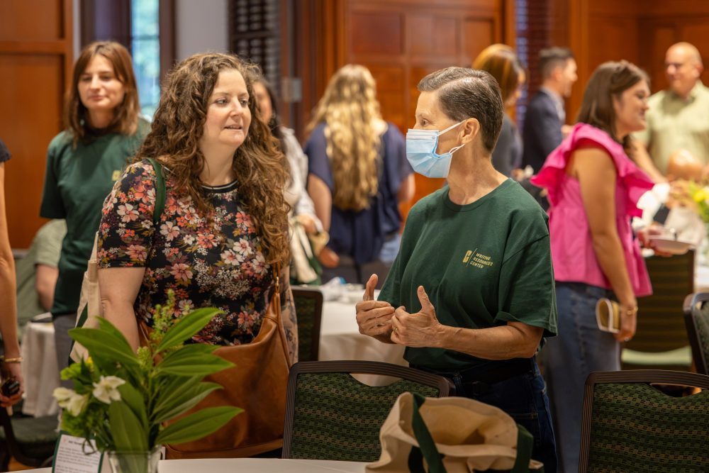Two individuals engage in conversation at a social gathering, with one wearing a face mask and both dressed in green shirts.
