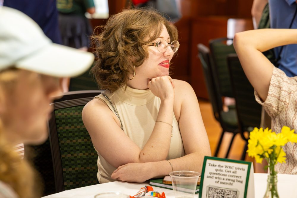 A person with curly hair and glasses sits at a table, resting their chin on their hand while looking thoughtfully to the side.